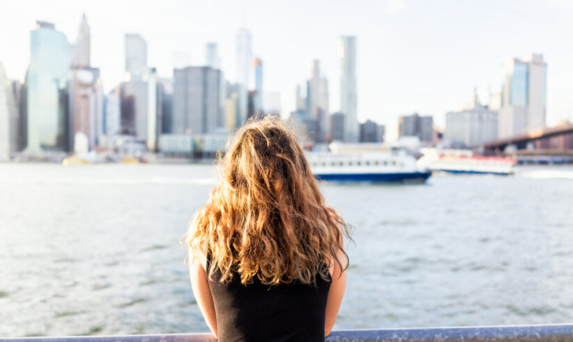 A person with curly hair, seen from behind, stands by a railing overlooking a river with a city skyline and a boat in the background on a sunny day.