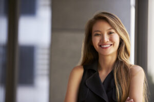 A young woman with long, light brown hair smiles confidently, standing with arms crossed in a sleeveless black top, in a modern indoor setting with blurred background.