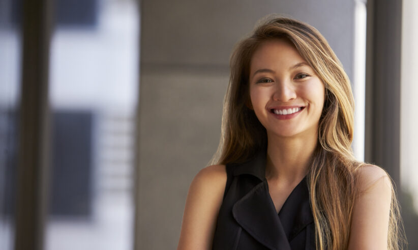 A young woman with long, light brown hair smiles confidently, standing with arms crossed in a sleeveless black top, in a modern indoor setting with blurred background.