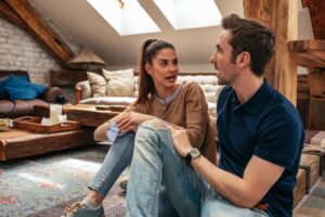 A young woman and man sit on the floor in a cozy, rustic living room, engaged in a lively conversation. The room has wooden beams, skylights, and casual decor with a tray and cup on the coffee table.