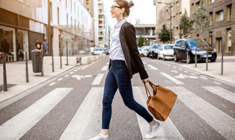 A woman with glasses, wearing a black blazer, white shirt, jeans, and white sneakers, walks across a crosswalk in an urban street, carrying a large brown handbag and smiling.