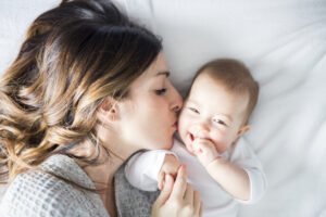 A woman lies on a bed, gently kissing a smiling baby on the cheek. The baby is wearing a white outfit and looks happy, holding the womans hand. Both are resting on white bedding.