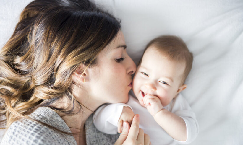 A woman lies on a bed, gently kissing a smiling baby on the cheek. The baby is wearing a white outfit and looks happy, holding the womans hand. Both are resting on white bedding.