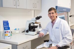A man in a white lab coat sits at a laboratory desk next to a microscope, test tubes, and medical supplies, smiling at the camera inside a bright, modern laboratory.