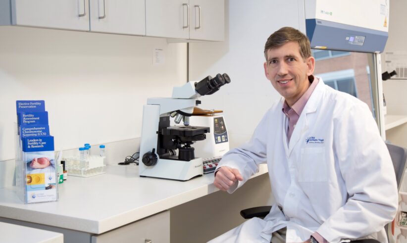 A man in a white lab coat sits at a laboratory desk next to a microscope, test tubes, and medical supplies, smiling at the camera inside a bright, modern laboratory.