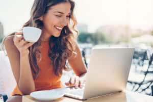 A smiling woman in an orange top sits outdoors, holding a cup of coffee in one hand and typing on a laptop with the other. A white saucer is on the table beside her. The background is bright and sunny.