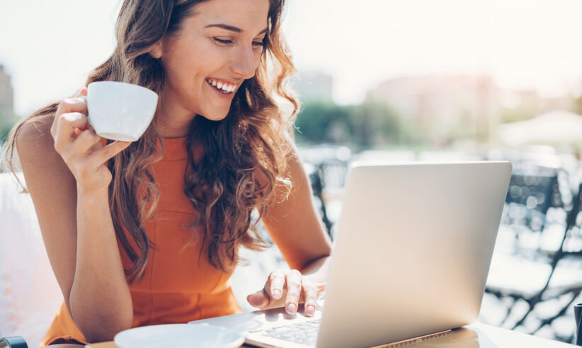 A smiling woman in an orange top sits outdoors, holding a cup of coffee in one hand and typing on a laptop with the other. A white saucer is on the table beside her. The background is bright and sunny.