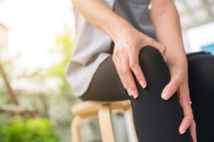 A person sitting on a wooden stool holds their knee with both hands, appearing to experience knee pain or discomfort. The background is softly blurred with natural light.
