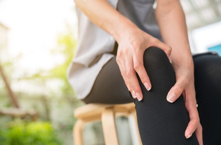A person sitting on a wooden stool holds their knee with both hands, appearing to experience knee pain or discomfort. The background is softly blurred with natural light.