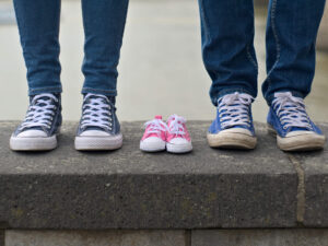Two adults in jeans and sneakers stand on either side of a small pair of pink baby shoes, placed between them on a stone ledge, suggesting an upcoming addition to the family.