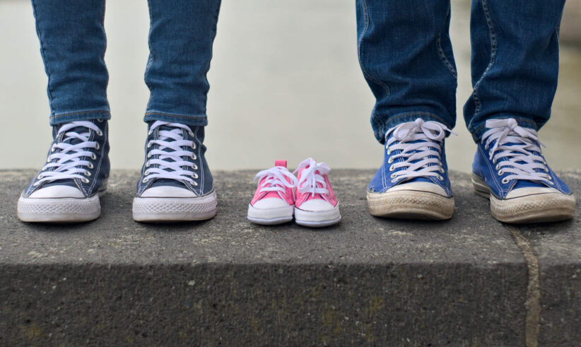 Two adults in jeans and sneakers stand on either side of a small pair of pink baby shoes, placed between them on a stone ledge, suggesting an upcoming addition to the family.