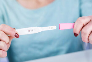 A person holding a pregnancy test with a single line in the result window, indicating a negative result. The person is wearing a light blue top and has red nail polish.