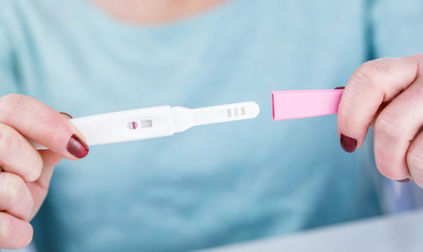 A person holding a pregnancy test with a single line in the result window, indicating a negative result. The person is wearing a light blue top and has red nail polish.