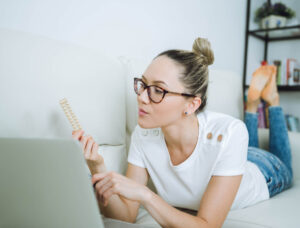 A woman wearing glasses lies on a couch while looking at a pack of birth control pills in one hand and using a laptop with the other.