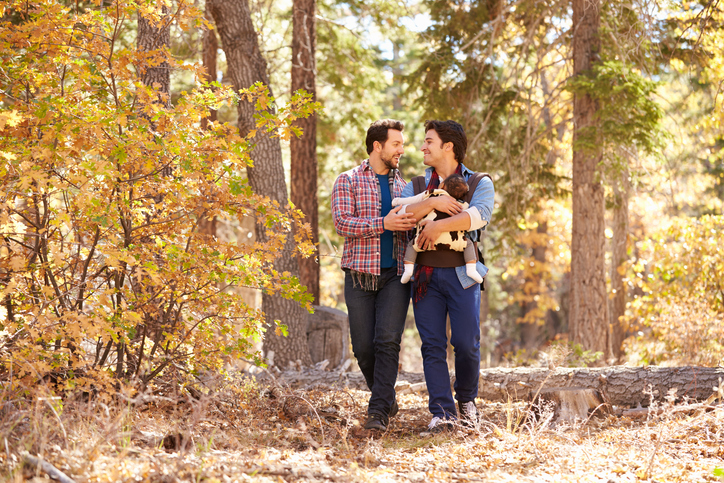 Two men walk through a sunlit forest, smiling at each other. One man is holding a baby in a carrier. Autumn leaves and tall trees surround them, creating a warm, natural setting.