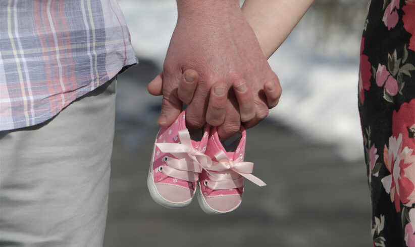 A couple holds hands while grasping a pair of small pink baby shoes, symbolizing an upcoming addition to the family. The man wears plaid, and the woman wears a floral dress.