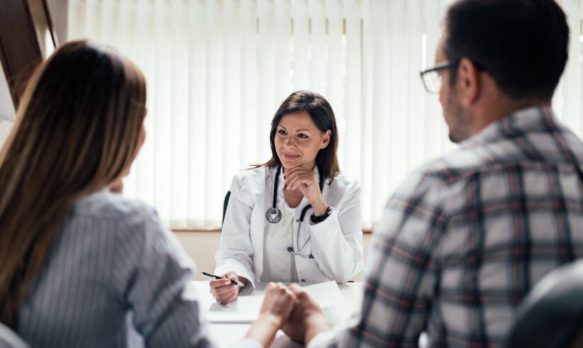A doctor in a white coat sits at a desk, smiling and talking with a couple who are holding hands. The doctor appears to be giving advice or consultation in a bright office with vertical blinds.