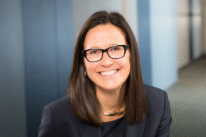 A woman with straight brown hair and black glasses smiles at the camera, wearing a dark blazer and a silver necklace, with a blurred office background.