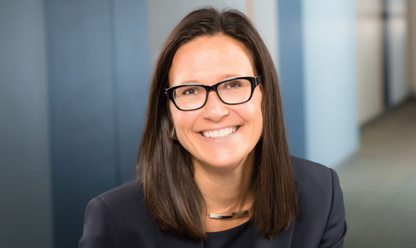 A woman with straight brown hair and black glasses smiles at the camera, wearing a dark blazer and a silver necklace, with a blurred office background.