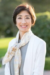 A woman with short brown hair smiles outdoors. She is wearing a white jacket, a grey top, and a patterned scarf tied around her neck. The background is blurred greenery and sunlight.