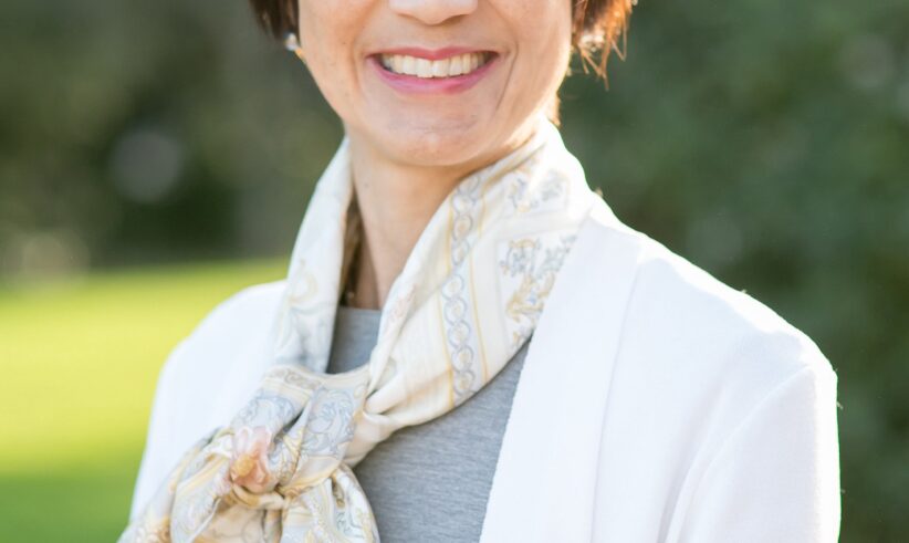 A woman with short brown hair smiles outdoors. She is wearing a white jacket, a grey top, and a patterned scarf tied around her neck. The background is blurred greenery and sunlight.