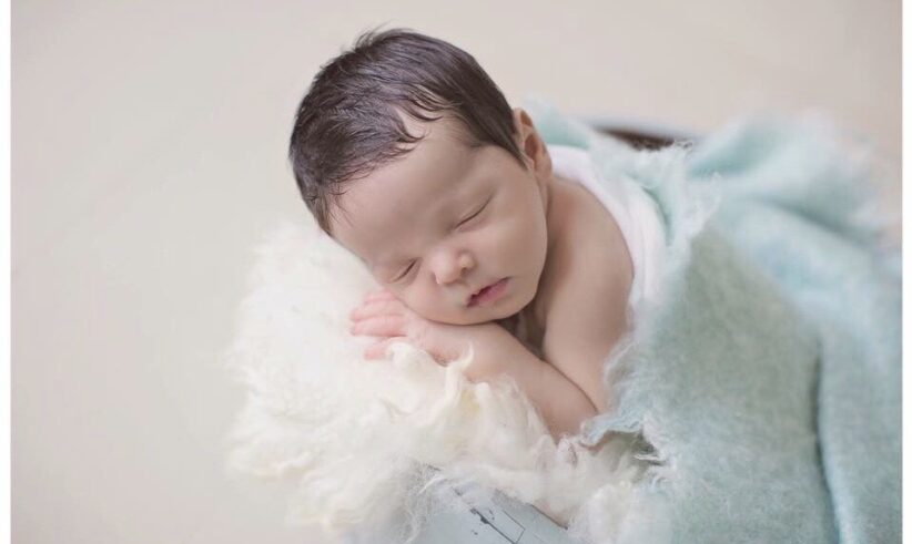 A newborn baby with dark hair sleeps peacefully on a soft white blanket, partially covered with a light blue blanket. The baby is resting on their side with their hands under their cheek.