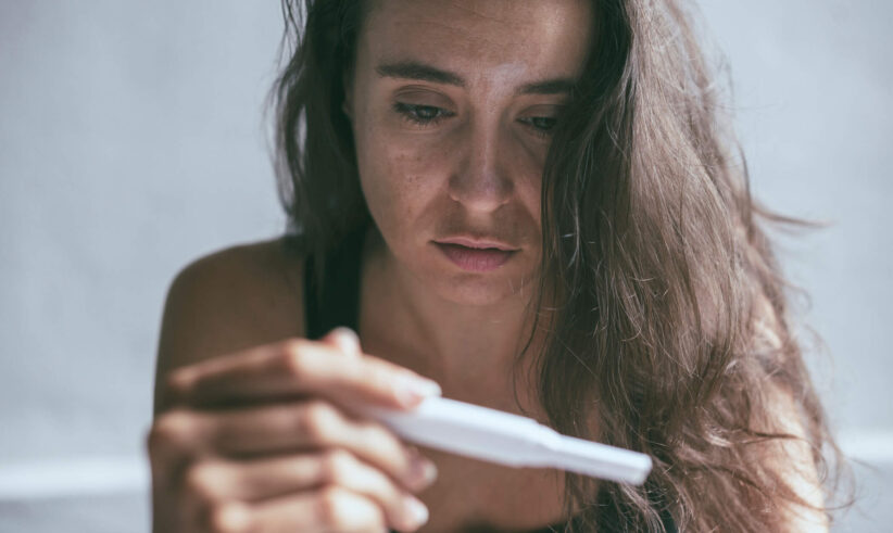 A woman with long brown hair looks intently at a pregnancy test she is holding, her expression appearing serious and contemplative.