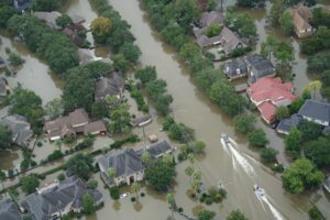 Aerial view of a flooded residential neighborhood with streets submerged, houses surrounded by water, and two boats navigating through the flooded area.