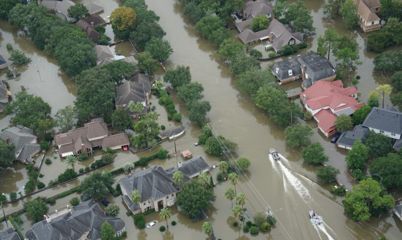 Aerial view of a flooded residential neighborhood with streets submerged, houses surrounded by water, and two boats navigating through the flooded area.