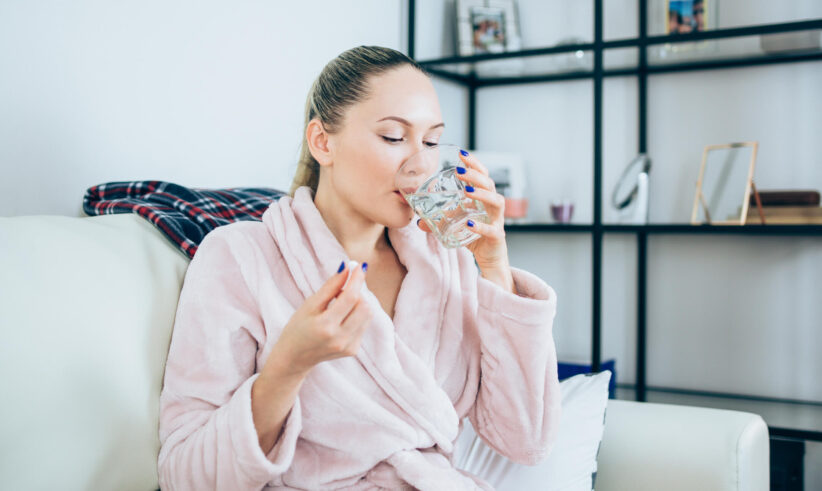 A woman in a pink bathrobe sits on a couch, holding a pill in one hand and drinking a glass of water. She appears relaxed in a cozy home setting with shelves and framed photos in the background.
