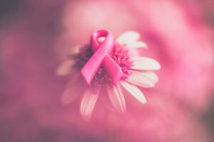 A pink breast cancer awareness ribbon rests on the center of a daisy flower, with a soft pink blurred background creating a gentle and supportive atmosphere.