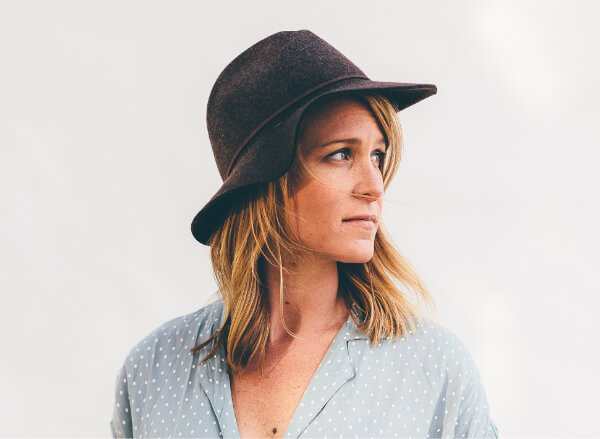 A woman with shoulder-length blonde hair wears a wide-brimmed brown hat and a light blue blouse with white polka dots, looking to her left against a plain light background.