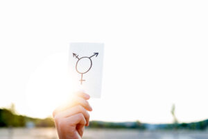 A hand holds up a white card with a transgender symbol drawn on it, against a bright, sunlit outdoor background.