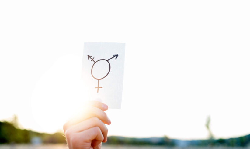 A hand holds up a white card with a transgender symbol drawn on it, against a bright, sunlit outdoor background.