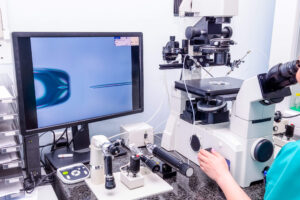 A laboratory scene shows a scientist using a microscope and micromanipulation equipment, with a monitor displaying a close-up of a cell and a needle, likely for in vitro fertilization or another microinjection procedure.