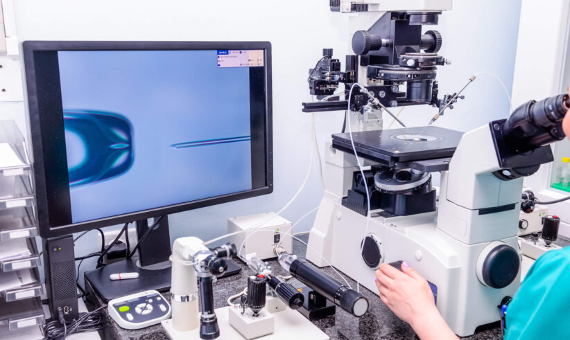 A laboratory scene shows a scientist using a microscope and micromanipulation equipment, with a monitor displaying a close-up of a cell and a needle, likely for in vitro fertilization or another microinjection procedure.