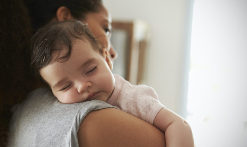 A baby sleeps peacefully on an adult’s shoulder, with the adult facing away and gently holding the child in a cozy, softly lit room.