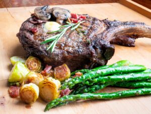 A grilled ribeye steak topped with mushrooms and rosemary sits on a wooden board, served with roasted Brussels sprouts, bacon pieces, and sautéed asparagus.