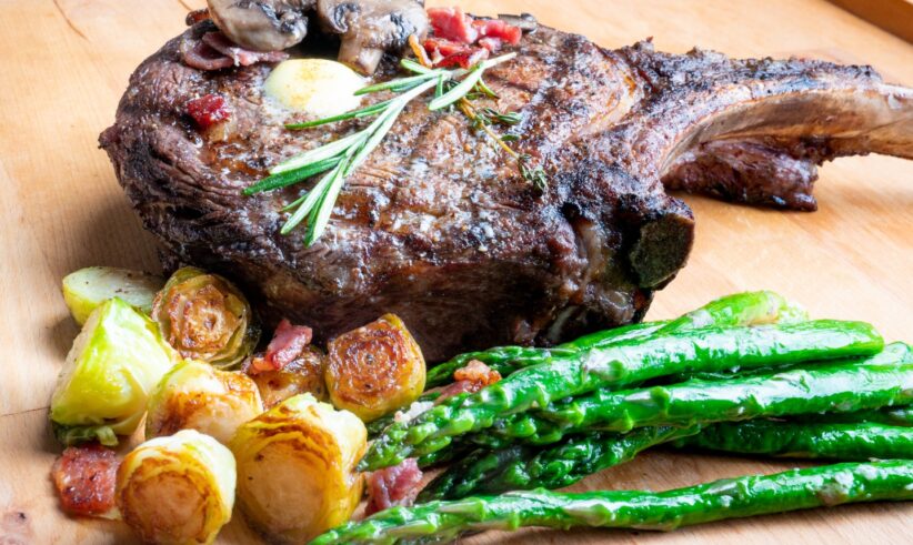 A grilled ribeye steak topped with mushrooms and rosemary sits on a wooden board, served with roasted Brussels sprouts, bacon pieces, and sautéed asparagus.