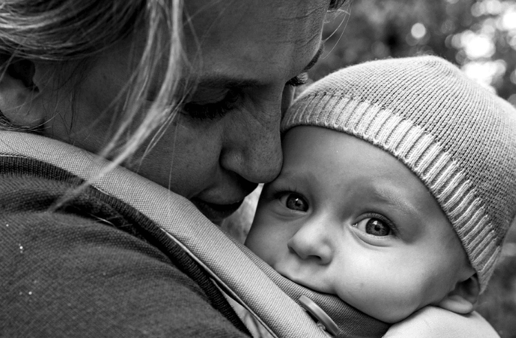 A close-up black and white photo of an adult lovingly holding a baby, who is wearing a knit hat and looking at the camera while nestled in a carrier. The adult’s face is close, showing affection and tenderness.