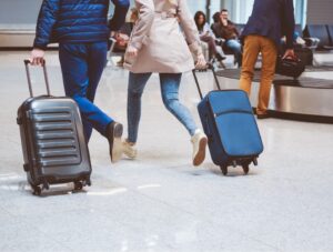 Two people walk quickly with rolling suitcases through an airport baggage claim area. Other travelers are sitting and standing in the background, and luggage carousels are visible.