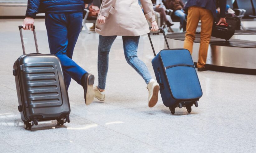 Two people walk quickly with rolling suitcases through an airport baggage claim area. Other travelers are sitting and standing in the background, and luggage carousels are visible.