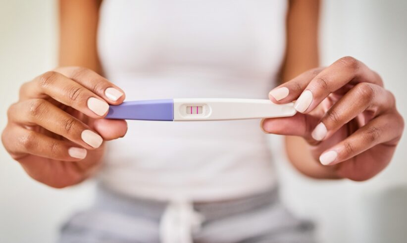 A person in casual clothes holds a pregnancy test showing two pink lines, indicating a positive result. The image focuses on the test and their hands.