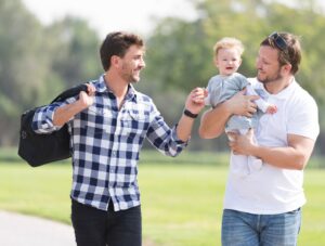 Two men stand outside on grass, smiling at each other. One is holding a baby, and the other carries a bag over his shoulder. The baby is also smiling. Trees and a blurred park background are visible.