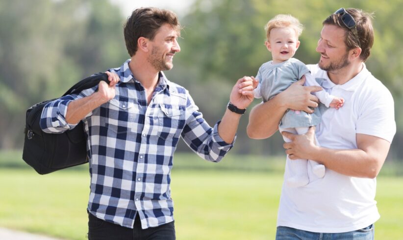 Two men stand outside on grass, smiling at each other. One is holding a baby, and the other carries a bag over his shoulder. The baby is also smiling. Trees and a blurred park background are visible.