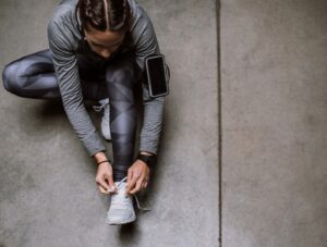 A person in athletic wear sits on the ground tying their shoe. They have a smartphone strapped to their upper arm and wear a smartwatch. The background is a smooth concrete floor.