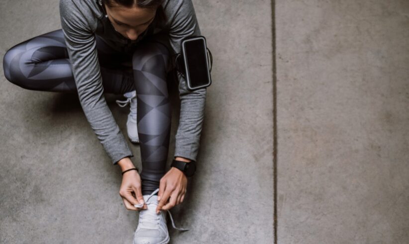 A person in athletic wear sits on the ground tying their shoe. They have a smartphone strapped to their upper arm and wear a smartwatch. The background is a smooth concrete floor.