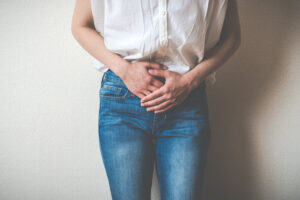 A person wearing a white shirt and blue jeans stands against a wall, holding their lower abdomen with both hands, suggesting discomfort or abdominal pain.