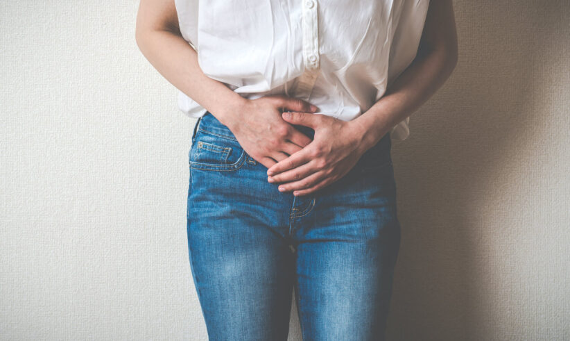 A person wearing a white shirt and blue jeans stands against a wall, holding their lower abdomen with both hands, suggesting discomfort or abdominal pain.