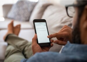 A person sitting on a couch uses a smartphone, browsing a website or app. The individual holds the phone in one hand and interacts with the screen using the other, with legs crossed and a relaxed posture.